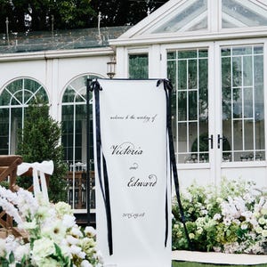 K&ouml;nnte beinhalten: Ein wei&szlig;es Hochzeits-Willkommensschild mit schwarzem Text und B&auml;ndern. Auf dem Schild steht "welcome to the wedding of Victoria and Edward 2025.09.08". Es wird im Freien vor einem wei&szlig;en Geb&auml;ude mit Glasfenstern und -t&uuml;ren aufgestellt.
