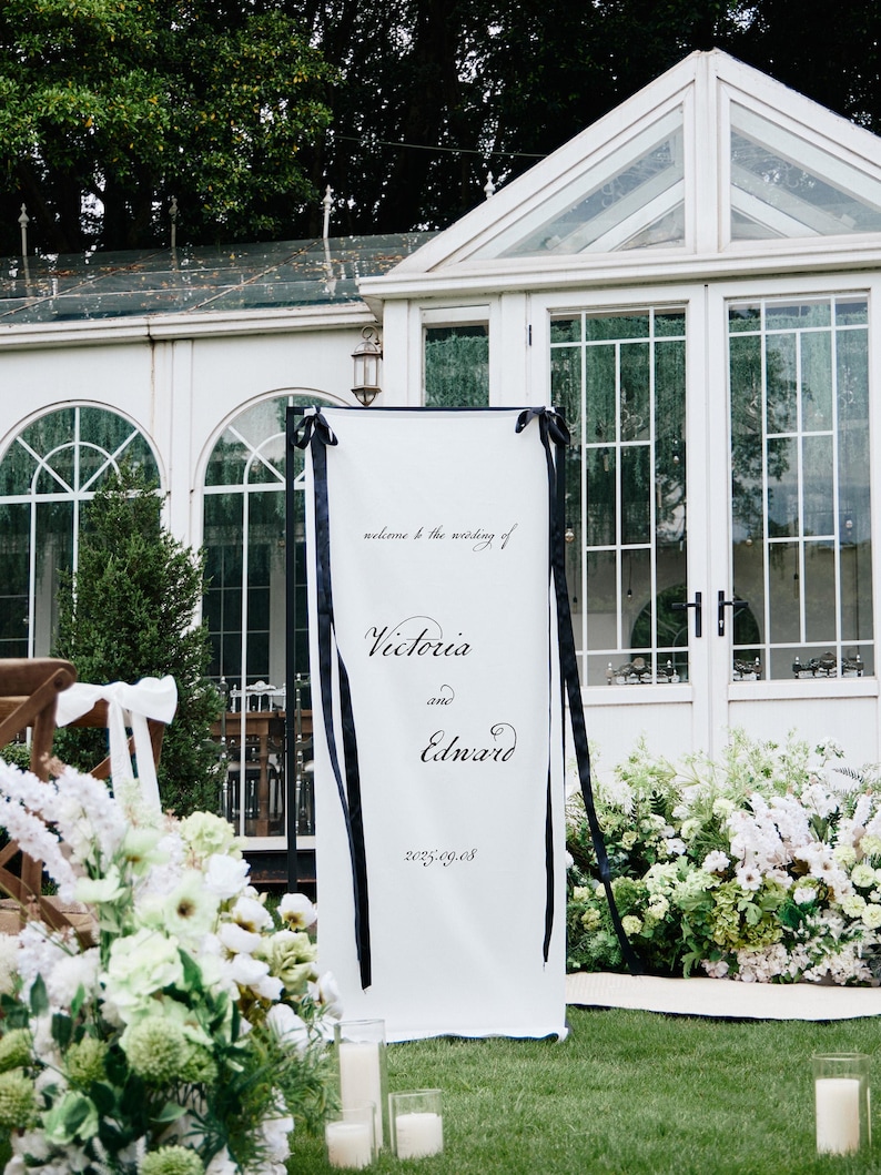 K&ouml;nnte beinhalten: Ein wei&szlig;es Hochzeits-Willkommensschild mit schwarzem Text und B&auml;ndern. Auf dem Schild steht "welcome to the wedding of Victoria and Edward 2025.09.08". Es wird im Freien vor einem wei&szlig;en Geb&auml;ude mit Glasfenstern und -t&uuml;ren aufgestellt.