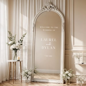 May include: A large, ornate, white-framed mirror with the text "WELCOME TO THE WEDDING OF LAUREL and DYLAN * MAY 2026". The mirror is surrounded by floral arrangements and a small side table with a vase and candle.