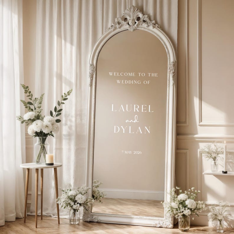May include: A large, ornate, white-framed mirror with the text "WELCOME TO THE WEDDING OF LAUREL and DYLAN * MAY 2026". The mirror is surrounded by floral arrangements and a small side table with a vase and candle.