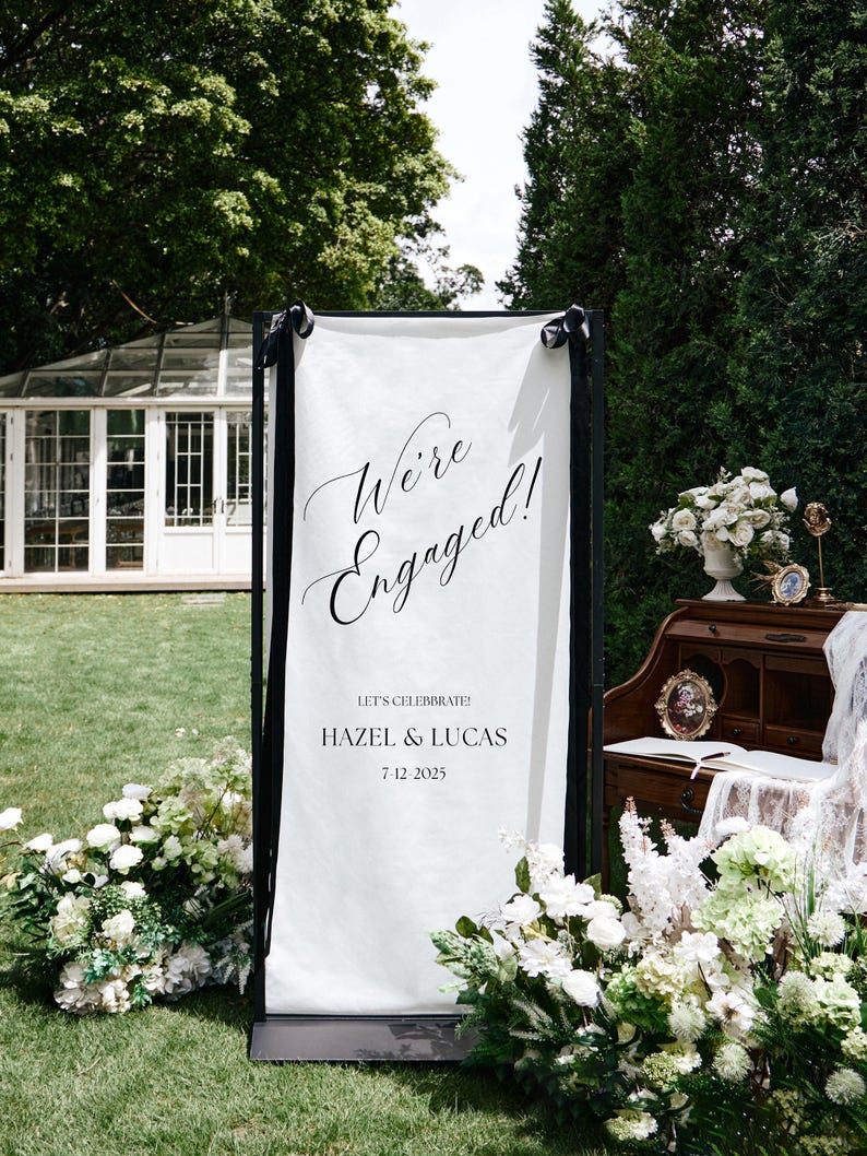 K&ouml;nnte beinhalten: Ein wei&szlig;es Verlobungsank&uuml;ndigungsbanner mit schwarzem Rahmen, auf dem die Worte "We're Engaged!" in Schreibschrift stehen. Die Namen "Hazel & Lucas" und das Datum "7-12-2025" sind ebenfalls zu sehen. Das Schild ist mit wei&szlig;en Blumen geschm&uuml;ckt.