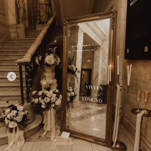 May include: A large, ornate gold-framed mirror with the words "welcome to the wedding of Vivian and Edmund 03.09.2026" etched on the surface. The mirror is surrounded by floral arrangements and candles, creating an elegant wedding decor.