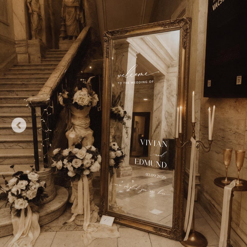 May include: A large, ornate gold-framed mirror with the words "welcome to the wedding of Vivian and Edmund 03.09.2026" etched on the surface. The mirror is surrounded by floral arrangements and candles, creating an elegant wedding decor.