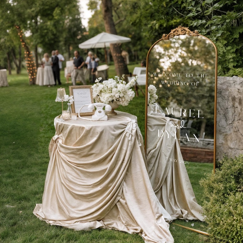 May include: An outdoor wedding reception scene featuring a table draped in champagne-colored fabric, adorned with a vintage telephone, flowers, and a framed sign. A large gold-framed mirror displays the text "Welcome to the Wedding of Laurel and Dylan, May 2026."