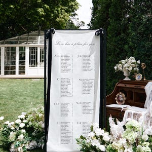 K&ouml;nnte beinhalten: Ein Hochzeits-Sitzplan auf einem schwarzen St&auml;nder mit wei&szlig;em Hintergrund. Die Namen sind alphabetisch aufgelistet. Der Text "Love has a place for you" steht oben. Wei&szlig;e Blumen und Gr&uuml;n umgeben den Plan.