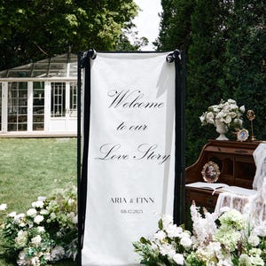 K&ouml;nnte beinhalten: Ein wei&szlig;es Willkommensschild mit schwarzem Rahmen, auf dem die Worte "Welcome to our Love Story" in eleganter Schrift stehen. Das Schild enth&auml;lt auch die Namen "Aria & Finn" und das Datum 08.12.2025. Wei&szlig;e Blumen umgeben das Schild.