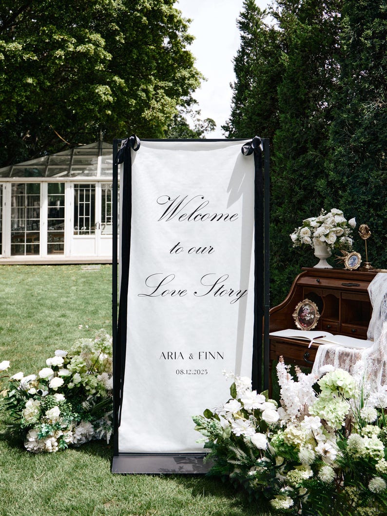 K&ouml;nnte beinhalten: Ein wei&szlig;es Willkommensschild mit schwarzem Rahmen, auf dem die Worte "Welcome to our Love Story" in eleganter Schrift stehen. Das Schild enth&auml;lt auch die Namen "Aria & Finn" und das Datum 08.12.2025. Wei&szlig;e Blumen umgeben das Schild.