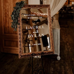 May include: A large, ornate mirror with a gold frame, displaying the text "WELCOME TO THE WEDDING OF LAUREL and DYLAN 7 MAY 2026." The mirror is propped on a black metal stand, with greenery draped on the side. The background shows a rustic wooden interior.