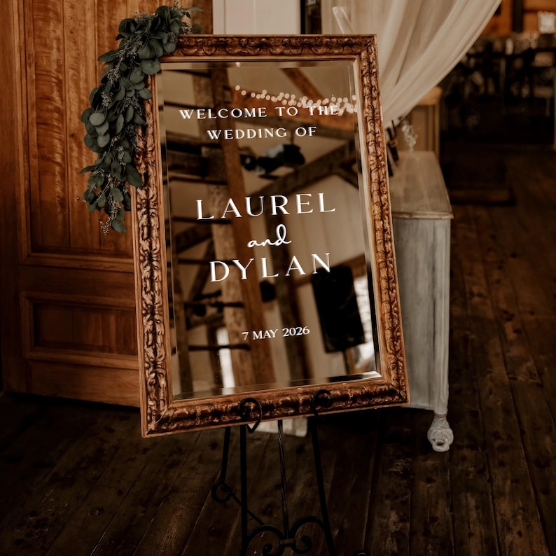 May include: A large, ornate mirror with a gold frame, displaying the text "WELCOME TO THE WEDDING OF LAUREL and DYLAN 7 MAY 2026." The mirror is propped on a black metal stand, with greenery draped on the side. The background shows a rustic wooden interior.