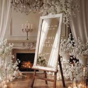 May include: A white framed mirror on a wooden easel, with the text "Welcome to the Wedding of Aurel and Dylan, 7 May 2026." The mirror is surrounded by white flowers, candles, and a chandelier, creating an elegant wedding scene.