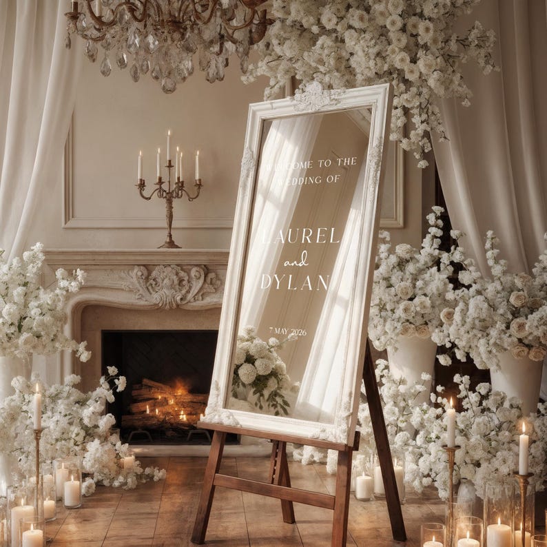 May include: A white framed mirror on a wooden easel, with the text "Welcome to the Wedding of Aurel and Dylan, 7 May 2026." The mirror is surrounded by white flowers, candles, and a chandelier, creating an elegant wedding scene.