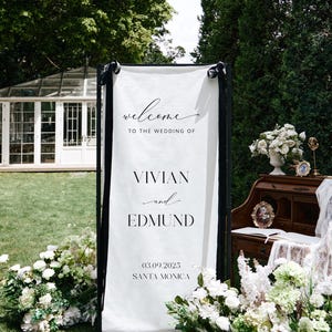 K&ouml;nnte beinhalten: Ein wei&szlig;es Hochzeits-Willkommensschild mit schwarzem Text: &bdquo;Willkommen zur Hochzeit von Vivian und Edmund 03.09.2025 Santa Monica&ldquo;. Das Schild ist schwarz umrahmt und mit schwarzen Schleifen verziert. Wei&szlig;e Blumen umgeben das Schild.