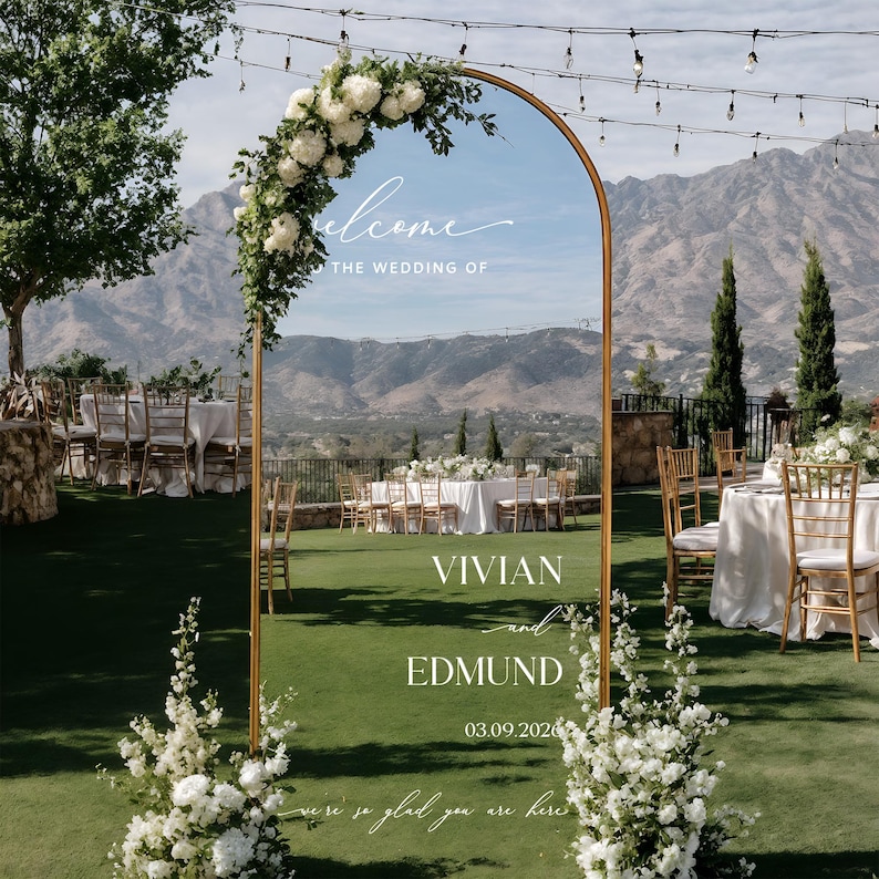 May include: A gold arched wedding sign with the names Vivian and Edmund, and the date 03.09.2026. The sign is decorated with white flowers and greenery, with the word "welcome" at the top. The sign is set up outdoors with a mountain backdrop.