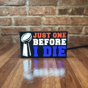May include: A black rectangular sign with the text "JUST ONE BEFORE I DIE" in orange, white, and blue. A football and trophy graphic is on the left. The sign is on a wooden surface.