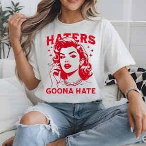 May include: White t-shirt with a red graphic of a woman smoking a cigarette, the text "HATERS GOONA HATE" is above and below the graphic. The shirt is worn with blue jeans.