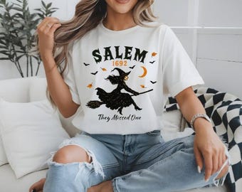 Puede incluir: Camiseta blanca con un gráfico de Salem 1692 que presenta una silueta de bruja en una escoba, una luna creciente y murciélagos. El texto "They Missed One" está debajo de la bruja. La camiseta es un diseño temático de Halloween.