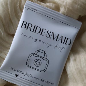 Puede incluir: Un paquete blanco con la inscripción "BRIDESMAID emergency kit" en negro, junto con un dibujo de un botiquín de primeros auxilios. El paquete también incluye el texto "FOR YOUR just in case MOMENTS" y un símbolo de corazón. La parte superior tiene una línea de puntos y la inscripción "open me".