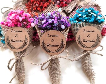Op de afbeelding: Kleine boeketten van kleurrijke gedroogde bloemen verpakt in jute. Elk boeket is vastgebonden met touw en heeft een rond label met de namen "Lorena & Ricardo" en een bladontwerp. De bloemen zijn roze, rood, paars en blauw.