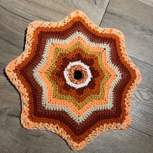 May include: A handmade, star-shaped crochet item in shades of orange, brown, and cream. The center is white with a dark hole. The edges are adorned with small, gold-colored beads. This decorative piece is likely a coaster or trivet.
