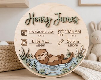 May include: A circular wooden birth announcement sign featuring the name Henry James in raised green lettering. It includes engraved details: November 2, 2024, 10:18 AM, 8 lbs 4 oz, and 20.5 in. A layered cutout of an otter swimming in water is at the base.