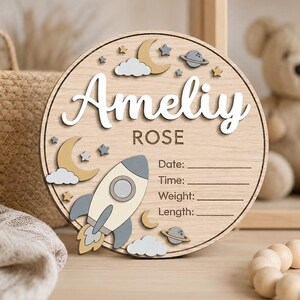Peut inclure: Un panneau de naissance circulaire en bois avec le nom Ameliy ROSE en texte blanc et marron. Le design comprend une fusée, des lunes, des étoiles, des nuages et des planètes, avec des lignes pour Date, Time, Weight et Length à compléter.