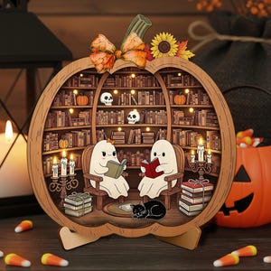 Könnte beinhalten: Holz-Kürbis-förmige Halloween-Dekoration mit einer Bibliotheksszene. Zwei Geisterfiguren lesen Bücher in Sesseln, mit einer schwarzen Katze. Die Bibliothek ist mit Büchern, Kürbissen und Totenköpfen gefüllt. Eine Schleife und eine Sonnenblume schmücken die Oberseite.