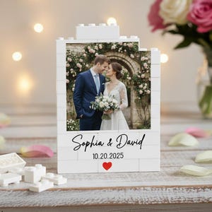 May include: A white brick-style photo frame featuring a wedding photo of a couple. The frame has the names "Sophia & David" and the date "10.20.2025" with a red heart. A bouquet of flowers is in the background.