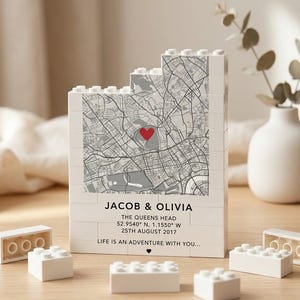 Puede incluir: Una pieza decorativa blanca en forma de bloque con un mapa y un corazón rojo. El texto dice "JACOB & OLIVIA", coordenadas y "LIFE IS AN ADVENTURE WITH YOU..." Varios bloques blancos están dispersos.