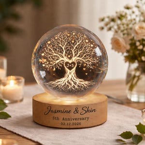 May include: A clear glass sphere with a tree of life design, illuminated from a wooden base. The base is engraved with the names "Jasmine & Shin", "5th Anniversary", and the date 02.12.2026. The sphere sits on a table with candles and flowers.