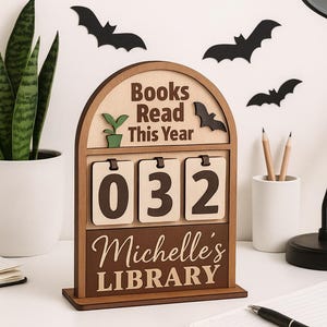 Könnte beinhalten: Ein Bücherzähler aus Holz mit dem Text "Books Read This Year" und "Michelle's LIBRARY". Der Zähler zeigt die Zahl "032" an. Dekorative Fledermäuse befinden sich an der Wand hinter dem Zähler. Eine Topfpflanze und Bleistifte sind ebenfalls sichtbar.