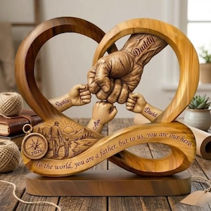 Könnte beinhalten: Holzskulptur mit herzförmigem Design und verschlungenen Händen. Die Skulptur enthält das Wort "Daddy" auf einer Hand und die Namen "Sophia", "Alex" und "David" auf anderen. Ein Kompass mit dem Jahr "2026" ist ebenfalls vorhanden, zusammen mit dem Text "To the world, you are a father, but to us, you are our hero."