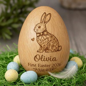 May include: A wooden Easter egg with a laser-etched design of a rabbit wearing a bow tie and floral details. The egg is engraved with the name "Olivia" and the text "First Easter 2026". The egg is surrounded by decorative Easter eggs and green grass.