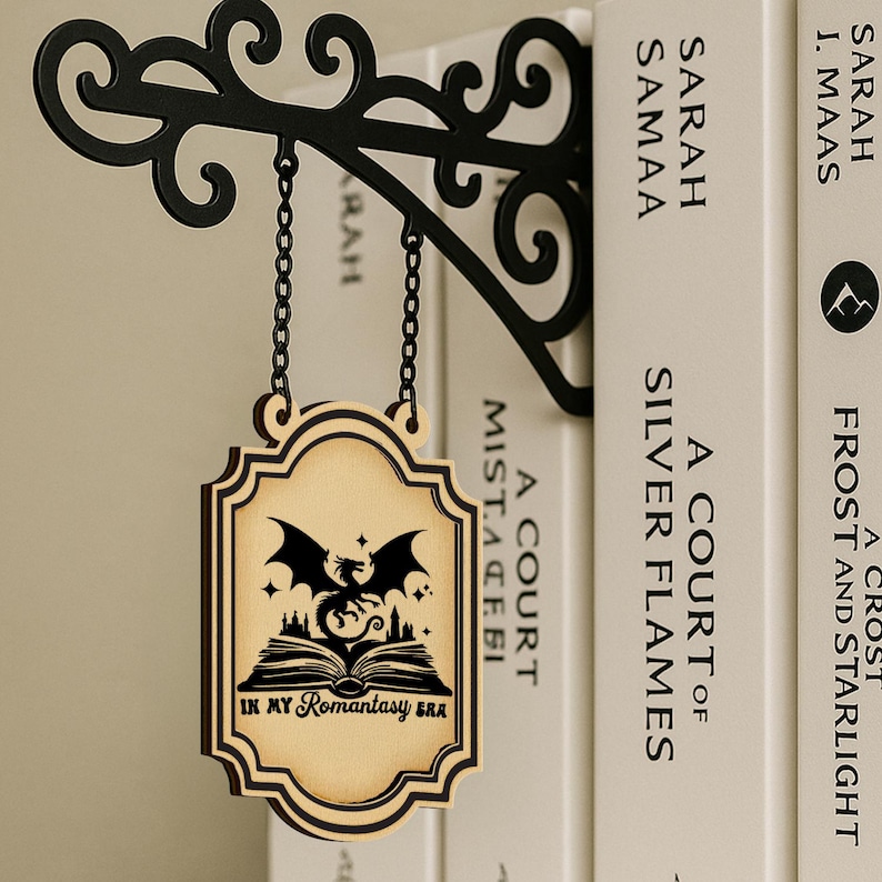May include: A wooden sign with a dragon and book design, hanging from a black metal bracket. The sign reads "IN MY Romantasy ERA". The bracket is attached to a row of books with white covers.