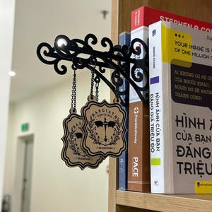 May include: Black metal bookshelf sign with ornate scrollwork and two hanging wooden plaques. The plaques feature a crest design with the text "BASGIATH WAR COLLEGE" and "DRAGON RIDER". The sign is attached to a bookshelf with books.