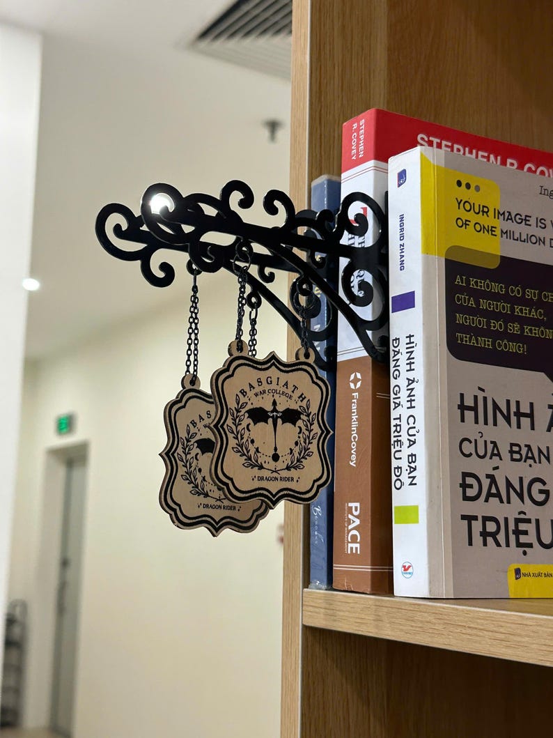 May include: Black metal bookshelf sign with ornate scrollwork and two hanging wooden plaques. The plaques feature a crest design with the text "BASGIATH WAR COLLEGE" and "DRAGON RIDER". The sign is attached to a bookshelf with books.