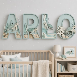 Puede incluir: Un conjunto de cuatro letras de pared dimensionales de color azul claro que forman la palabra ARLO. Cada letra presenta recortes en capas de tortugas marinas, corales y conchas en tonos verde, beige y blanco.