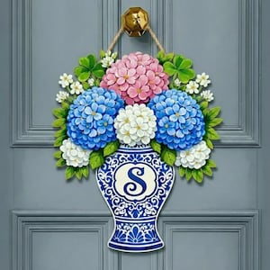May include: A wooden door hanger shaped like a vase filled with pink, blue, and white hydrangea flowers. The vase is blue and white with the letter "S" in the center. It hangs from a gold-colored door knocker.
