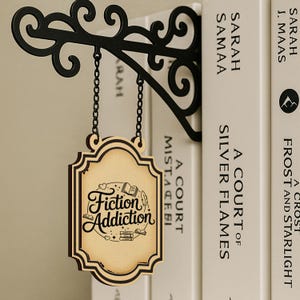 以下が含まれることがあります： 黒い金属製のブラケットから吊り下げられた、「Fiction Addiction」と書かれた木製の看板。看板は薄茶色の背景に濃い茶色の縁取りがあり、本と羽のイラストが描かれています。背景には本が見えます。