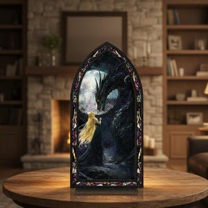 Op de afbeelding: Een gotisch gebrandschilderd glazen paneel met een draak en een vrouw met lang blond haar. Het kunstwerk staat tegen een nachtelijke hemel met een volle maan. Het paneel is zwart omlijst met paarse accenten en staat op een houten tafel.