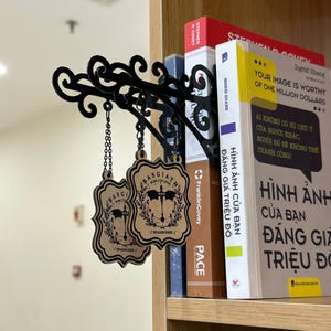 May include: Black metal decorative shelf bracket with ornate scrollwork, holding two wooden Basgiath War College signs. The signs are light brown with black text and a crest design, hanging by chains. Books are visible on the shelf.