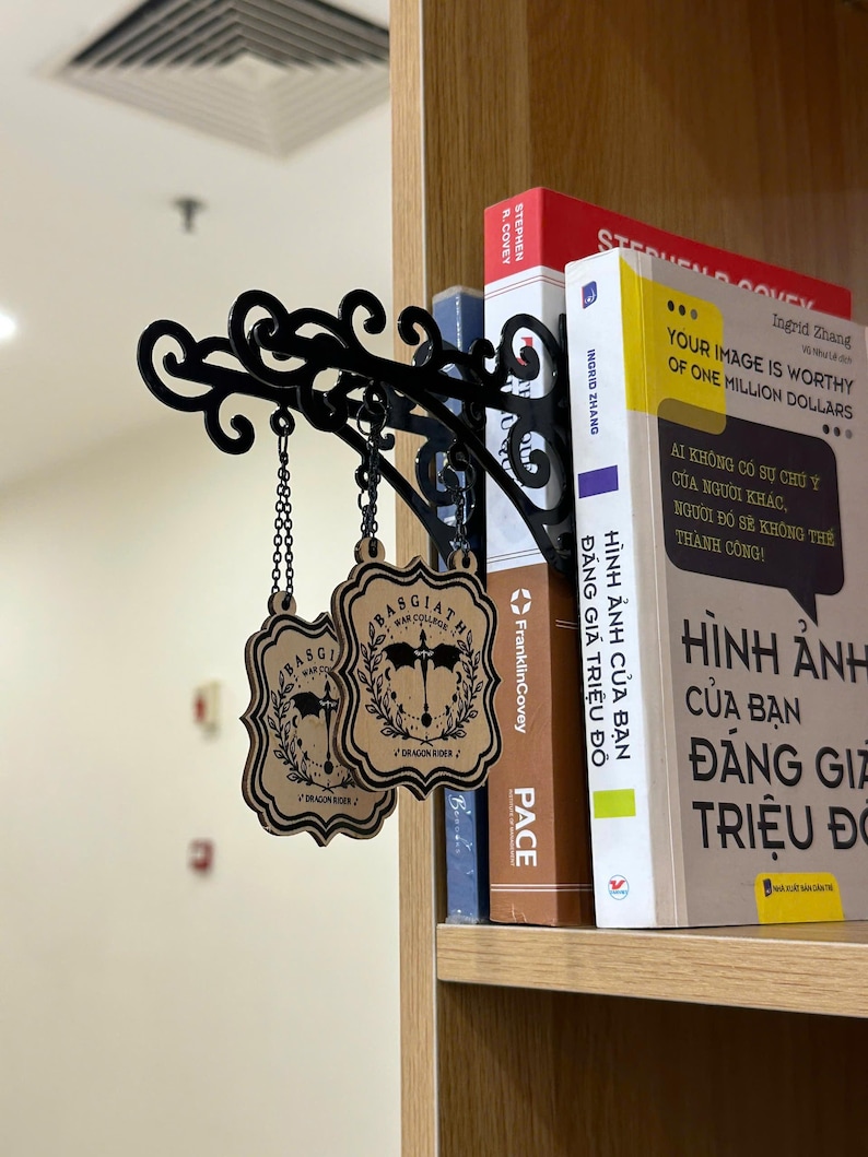 May include: Black metal decorative shelf bracket with ornate scrollwork, holding two wooden Basgiath War College signs. The signs are light brown with black text and a crest design, hanging by chains. Books are visible on the shelf.