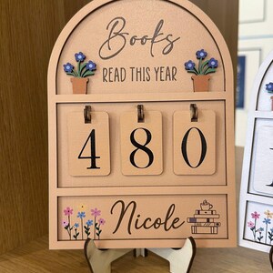 May include: A wooden book counter displaying "Books Read This Year" and the number "480". The counter features a floral design and the name "Nicole". It is supported by a wooden stand. The design is in a light brown color.