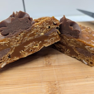 May include: Two pieces of fudge on a wooden board. The fudge has layers of caramel, chocolate, and a crunchy base. Each piece is topped with a dollop of chocolate frosting. A knife is visible in the background.