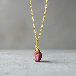 May include: A gold-colored chain necklace with a pendant featuring a raw, uncut ruby gemstone. The ruby is a deep pink color with some green and brown inclusions. The necklace is displayed against a gray background.