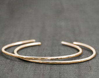 May include: Two gold-toned cuff bracelets with a hammered, textured finish. Each bracelet features a thin, open-ended circular band design with rounded ends. The metal surface reflects light, highlighting the irregular, dimpled pattern across the exterior.