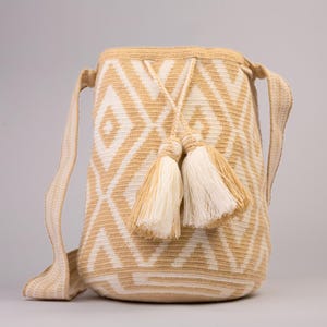 Op de afbeelding: Een beige en witte geweven schoudertas met een geometrisch ruitpatroon. De tas heeft een trekkoordsluiting en twee decoratieve kwastjes. De schouderriem is ook geweven. De tas is ongeveer 30 cm hoog en 25 cm breed.
