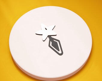 May include: A white star-shaped object with a black diamond-shaped tail, resting on a light pink circular base. The background is a bright yellow. The star and diamond are likely decorative elements.