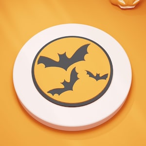 Peut inclure: Un dessous de verre rond blanc sur le thème d'Halloween avec un centre jaune représentant trois silhouettes de chauves-souris noires. Le dessous de verre est posé sur un fond orange, créant un design festif et saisonnier.