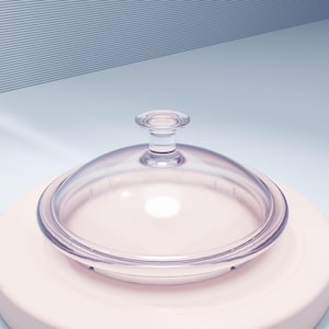Könnte beinhalten: Ein klarer Glasdeckel mit runder Form und kleinem Griff oben. Der Deckel befindet sich auf einer hellrosa Oberfläche, mit einem hellblauen und weißen Hintergrund.
