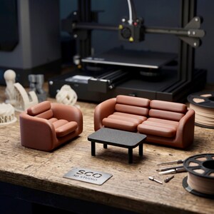 May include: A miniature brown sofa and matching armchair with a black rectangular coffee table. A metal plate with the text SCO 3D STUDIO is visible. The furniture set is displayed on a wooden surface with 3D printing equipment in the background.